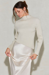 A woman is wearing a high-neck, long-sleeved ribbed sweater in a light, creamy color. She pairs it with a silky, satin-like skirt in an ivory shade that has a smooth, shiny texture.
